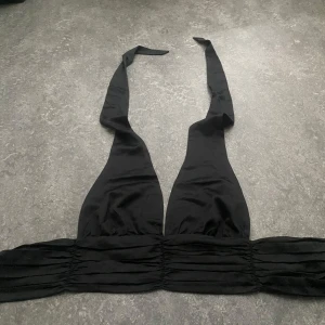 Svart halterneck topp från Bik Bok - Snygg svart halterneck topp från Bik Bok i slätt, glansigt tyg. Toppen har bred smockad midja, djup v-ringning och knytband i nacken. Perfekt för att skapa en cool och trendig look.