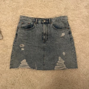 Blå jeanskjol med slitningar - Snygg blå jeanskjol med rå kant och slitna detaljer framtill. Klassisk femficksmodell med dragkedja och knapp i midjan. Perfekt för dig som gillar en avslappnad och trendig stil.