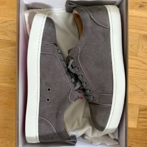 Grå mockasneakers från Louboutin - Snygga grå sneakers i mocka från Louboutin med klassisk vit sula och ikonisk röd undersida. Skorna har rund tå, snörning och stilren design som passar till det mesta. Perfekta för dig som gillar exklusiva detaljer och vill sticka ut med en lyxig touch.