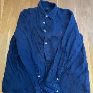 Blå linneskjorta från Polo Ralph Lauren - Snygg marinblå skjorta från Polo Ralph Lauren i 100% linne. Klassisk krage, långa ärmar och knäppning framtill med vita knappar. Ikoniska röda broderade loggan på bröstet ger en stilren touch. Perfekt för dig som gillar en avslappnad men ändå uppklädd look.