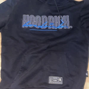 Svart hoodie från Hoodrich med tryck - Svart hoodie från Hoodrich med stort grått och blått tryck på bröstet. Klassisk känguruficka framtill, huva med snörning och en liten etikett nertill. Perfekt för en chill och street-inspirerad stil.