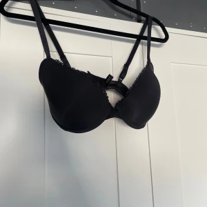 Svart push-up bh med spetsdetaljer - Svart push-up bh från twilfit med tunna axelband och fina spetsdetaljer längs kanten. Bh:n har vadderade kupor och klassisk knäppning bak. Resoren är lite trasig men inget som stör användandet.