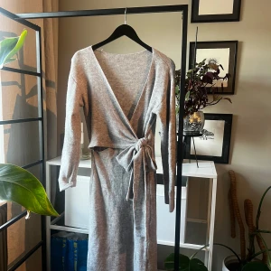 Grå omlottklänning i stickat material - Mysig grå lång stickad klänning med omlott och knyt i midjan. Klänningen har lång ärm och v-ringning framtill, vilket ger en avslappnad men ändå stilren look. Perfekt för dig som gillar bekväma och snygga outfits under kyligare dagar.