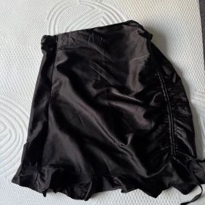 Svart silkeskjol med volang - Supersnygg svart kortkjol i satinliknande material med omlottdesign och volangkant nertill. Kjolen har en lätt glansig finish och dragsko på sidan för extra detalj. Perfekt för dig som gillar trendiga och feminina plagg.