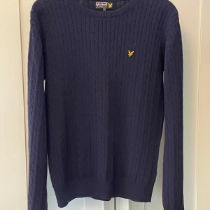 Marinblå stickad tröja Lyle & Scott - Säljer en marinblå kabelstickad tröja från Lyle & Scott med gul logga på bröstet. Tröjan har rund halsringning, långa ärmar och ribbade muddar. Perfekt för dig som gillar klassisk och stilren look.