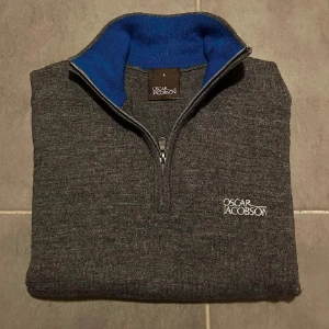 Grå half zip tröja Oscar Jacobson - Mörkgrå stickad tröja från Oscar Jacobson med halv dragkedja och broderad logga på bröstet. Insidan av kragen är blå för en snygg kontrast. Långärmad modell med ribbade muddar och nederkant. Perfekt för dig som gillar stilren och klassisk look. 