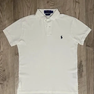 Vit pikétröja från Polo Ralph Lauren - Klassisk vit pikétröja från Polo Ralph Lauren med korta ärmar och blå broderad logga på bröstet. Tröjan har krage, knappar framtill och är tillverkad i mjuk bomull. Perfekt för en clean och stilren look.