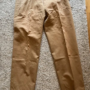 Louis Vuitton Beiga byxor  - Snygga beiga byxor från Louis Vuitton med exklusiv LV-detalj i guld vid midjan. Byxorna har rak passform och är tillverkade i ett mjukt bomullsmaterial. Perfekta för dig som vill ha en clean och lyxig look. Kemtvätt har gjort att del av fodret färgats. 