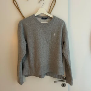 Grå sweatshirt från Polo Ralph Lauren - Klassisk grå sweatshirt från Polo Ralph Lauren med rund halsringning och broderad logga på bröstet. Tröjan har långa ärmar och ribbade muddar vid hals, ärmslut och nederkant. Perfekt för en chill och stilren look.