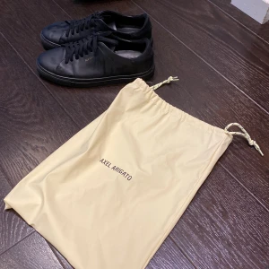 Svarta sneakers från Axel Arigato - Stilrena svarta sneakers från Axel Arigato i slät skinn med diskret guldfärgad logga på sidan. Klassisk låg modell med rund tå och svarta snören. Kommer med original dustbag. Perfekta för dig som gillar minimalistisk och clean stil. Använda fåtal gånger, ny pris=2500kr mitt pris 1500kr