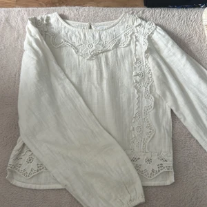 Vit blus med spetsdetaljer - Superfin vit blus med långa ärmar och snygga spetsdetaljer längs halsringning och sidor. Blusen har en lätt och luftig känsla, perfekt för vår och sommar. Den har en avslappnad passform och är tillverkad i ett tunt tyg som känns skönt mot huden. Har små fläckar som inte syns med tröjan på.