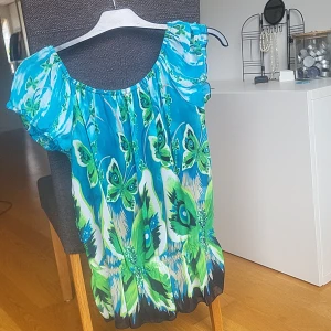 Blågrön blus med fjärilsmönster - Färgstark blus i blått och grönt med stora fjärilar och puffiga korta ärmar. Toppens material är tunt och flowy, troligen polyester, med resår vid halsringningen. Perfekt för dig som vill sticka ut med ett unikt mönster och somrig vibe.