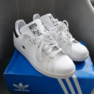 Adidas Stan Smith sneakers still 39 - Klassiska Adidas Stan Smith sneakers i vitt skinn med svarta detaljer på hälen och logga på plösen. Snygg minimalistisk design med rund tå, perforerade sidor och snörning. Perfekta för en clean och tidlös look. Använda bara en gång, är i mycket fint skick. Nypris 1400 kr