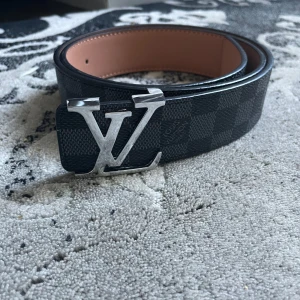 Bälte  rutigt skärp i skinn - Snyggt  bälte i svart och grått med klassiskt rutigt mönster i silvermetall. Bältet är gjort i skinn med ljusbrun insida och har exklusiv känsla. Perfekt accessoar för att lyfta din outfit.