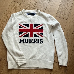 Vit stickad tröja Morris Union Jack - Vit stickad tröja från Morris med Union Jack-flagga och tydlig MORRIS-text framtill. Klassisk rund halsringning och ribbade muddar vid ärmslut och nederkant. Perfekt för dig som gillar preppy stil och vill sticka ut med snygg logga.