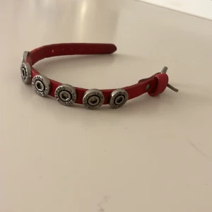 Rött läderarmband från Diesel - Coolt rött armband i läder från Diesel med silverfärgade metalldetaljer och nitar längs bandet. Armbandet har justerbart spänne och rundade dekorationer med Diesel-logga, perfekt för en edgy look.
