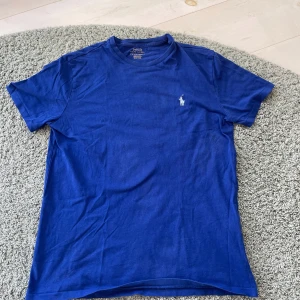 Blå t-shirt från Polo Ralph Lauren - Klassisk blå t-shirt från Polo Ralph Lauren med vit broderad logga på bröstet. T-shirten har rund halsringning och korta ärmar. Tillverkad i mjuk bomull som är skön mot huden. Perfekt för en clean och stilren look.