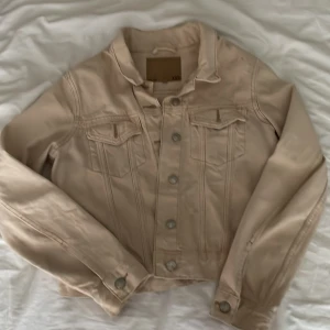 Beige jeansjacka från 157 - Snygg beige jeansjacka från 157 med klassisk krage, bröstfickor med lock och silverfärgade knappar. Jackan har raka sömmar och är tillverkad i bomull. Perfekt för dig som gillar en clean och stilren look.Finns någon fläck upptill som syns på bilderna men annars i mycket fint skick.