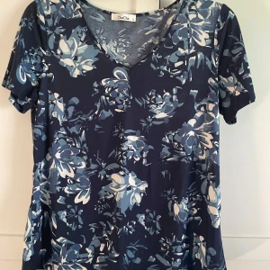 Blommig topp från ChaCha i blått - Snygg topp från ChaCha med blommigt mönster i olika blå nyanser och vitt. Modellen har v-ringning och korta ärmar. Materialet känns mjukt och följsamt, perfekt för dig som gillar en avslappnad men ändå stilren look.