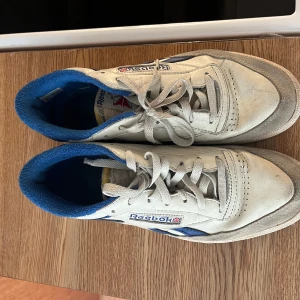 Vita och blå Reebok sneakers - Klassiska Reebok sneakers i vitt skinn med blå detaljer och blå insida. Modellen har snörning, platt sula och rund tå. Perfekta för dig som gillar retrostil och sportig vibe. Storlek 42,5 EU, med tydlig Reebok-logga på sidan. Nypris 1100, använd fåtal gånger 