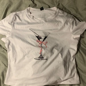 Vit t-shirt med martiniglas-tryck - Vit t-shirt i bomull med ett tryck av ett martiniglas och en rosa rosett framtill. Klassisk rund halsringning och korta ärmar. Perfekt för dig som gillar coola och lekfulla prints.❤️