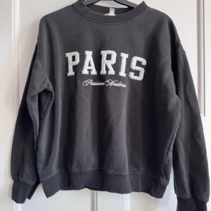 Svart sweatshirt med Paris-tryck - Cool svart sweatshirt med stort vitt PARIS-tryck och broderad text på bröstet. Tröjan har rund hals, ribbade muddar och är långärmad. Perfekt för dig som gillar streetstyle och vill ha en chill vibe.