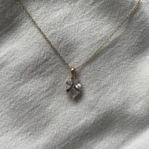 Edblad halsband - Ett elegant halsband med tunn guldfärgad kedja och ett hänge i form av en klar kristallslipning, infattad i guld. Hänget har en unik form med två små stenar ovanför en större fyrkantig kristall. Perfekt för dig som gillar stilrena smycken med lite extra bling.