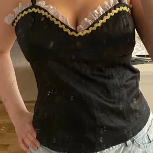 Svart korsett linne/ topp med spets och guld och detaljer - Svart korsett-topp med smala axelband med vit spets längs urringningen och en vågig guldfärgad kant. Sitter figurnära och har en feminin, edgy vibe. Perfekt för dig som vill sticka ut med detaljer och kontraster. Står strl L men kan sitta som M
