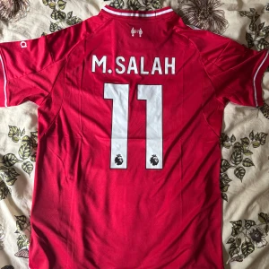 Liverpool Salah 11 Adidas matchtröja - Röd Liverpool fotbollströja med vita detaljer, Adidas-logga och klubbmärke på bröstet. Nummer 11 och M. Salah tryckt på ryggen. Klassisk design med vita ränder på axlarna och sponsortryck framtill. Tillverkad i lätt och ventilerande material.