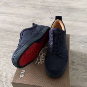 Mörkblå sneakers från Louboutin - Snygga mörkblå sneakers från Christian Louboutin i mocka med ikonisk röd sula. Skorna har snörning, rund tå och diskret broderad logga på hälen. Perfekta för dig som vill sticka ut med exklusiv stil och lyxiga detaljer.