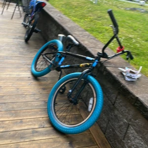 Bmx cykel  - Bilderna visar en BMX-cykel med blå däck och svarta detaljer, dekorerad med klistermärken. Cykeln ser robust ut och passar perfekt för dig som gillar tricks och street riding. Perfekt för unga som vill ha en cool och personlig cykel att sticka ut med på stan eller i skateparken.