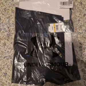 Svarta träningsbyxor från Under Armour - Svarta träningsbyxor från Under Armour med elastisk midja och snörning. Byxorna har färgglada detaljer och logga längs benet. Tillverkade i ett lätt och funktionellt syntetmaterial som passar perfekt för träning.