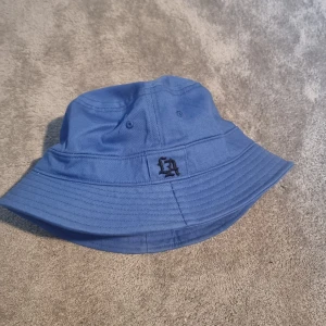 Blå LA bucket hat i bomull - Snygg blå bucket hat med svart LA-brodyr framtill. Klassisk form med bred, nedåtlutande brätte och sydda detaljer. Tillverkad i mjuk bomull som andas, perfekt för soliga dagar. Enkel och stilren design som lyfter vilken outfit som helst.