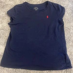 Marinblå t-shirt från Polo Ralph Lauren - En marinblå t-shirt från Polo Ralph Lauren med klassisk rund hals och korta ärmar. På bröstet finns den ikoniska röda Polo-loggan broderad. T-shirten är i mjuk bomull och har en enkel, stilren look som är lätt att matcha. Stoleken är 160. Man kan diskutera priset dessutom.