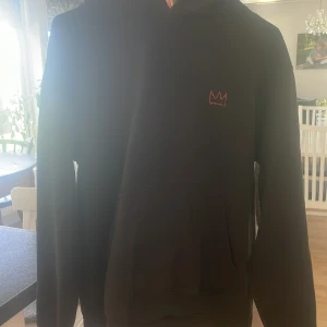 Svart HOV1 hoodie med tryck - Svart hoodie från MOV1 med röd krona broderad på bröstet och stort grafiskt tryck på ryggen med turnédatum och text. Klassisk känguruficka framtill och huva. Perfekt för dig som gillar streetwear och vill sticka ut.