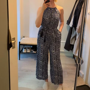 Blå mönstrad byxdress med knyt - Säljer en blå byxdress med blommigt och randigt mönster. Byxdressen har halterneck, knytning i midjan och öppen rygg med knytband. Lätt och luftig, perfekt för varma dagar. Vida ben och snygg passform.