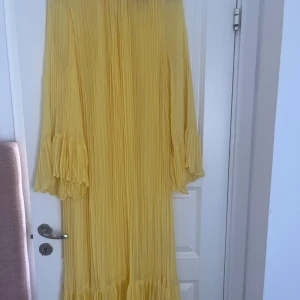 Gul långklänning med volangärmar XS - Lång gul klänning i plisserat tyg med vida, volangprydda ärmar och rund halsringning. Klänningen har en luftig och flowy känsla, perfekt för varma dagar. Stängs i nacken och har fina detaljer vid ärmslut och nederkant.
