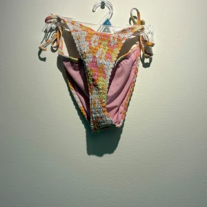 Färgglad bikini med smock och knyt - Superfin bikini i smockad modell med knytband i sidorna och baktill. Färgerna är en mix av vit, rosa, gul och orange i ett lekfullt mönster. Toppen är bandeau-stil och trosorna har hög midja. Perfekt för stranden eller poolen i sommar!