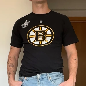 Boston Bruins t-shirt - Svart Boston Bruins t-shirt från Reebok i 100% bomull. Stor logga på bröstet, NHL-märke och Stanley Cup-detalj framtill. På ryggen står det THOMAS och nummer 30 i gult och vitt. Perfekt för hockeyfans som vill sticka ut.