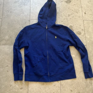 Blå hoodie från Polo Ralph Lauren - Säljer en snygg blå hoodie från Polo Ralph Lauren med dragkedja och det klassiska gula logotypbroderiet på bröstet. Perfekt för en avslappnad stil. Barn storlek men passar S, skicket är bra och ganska oanvänd