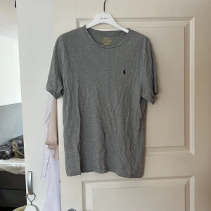 Grå t-shirt från Ralph Lauren - Säljer en klassisk grå t-shirt från Ralph Lauren med den ikoniska broderade loggan på bröstet. T-shirten är kortärmad och tillverkad i mjuk bomull. Perfekt för en avslappnad stil.