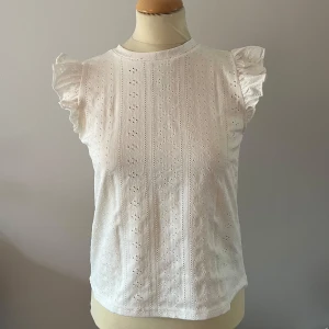 Vit blus med spetsdetaljer - Säljer en söt vit blus med spetsdetaljer och korta ärmar med volang. Perfekt för en somrig look. Blusen har ett diskret mönster och en rund halsringning.