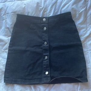 Svart jeanskjol från H&M - Snygg svart jeanskjol från H&M med knappar framtill. Kjolen har en klassisk design med fickor och är perfekt för en stilren look. Passar bra till olika tillfällen och kan enkelt matchas med olika toppar.