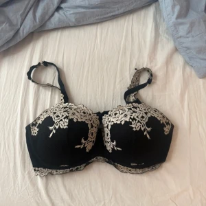 Svart spets-bh med broderade detaljer - Elegant svart bh med vackra broderade spetsdetaljer i vit. Justerbara axelband och hakknäppning bak för perfekt passform. Perfekt för en sofistikerad look. Ny pris: 500kr