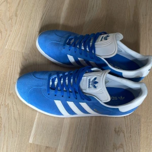 Blå Adidas Gazelle sneakers - Säljer ett par klassiska Adidas Gazelle sneakers i en snygg blå färg med vita detaljer. Skorna har den ikoniska tre-randiga designen och är tillverkade i mocka. Perfekta för en stilren och sportig look.