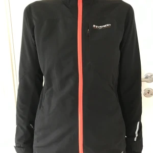 Svart träningsjacka Everest stl 36 Ny - Svart jacka Everest med orange dragkedja framtill. Stl 36. Jackan har en liten bröstficka med dragkedja och reflexdetaljer på ärmarna och ryggen. Två sidfickor med dragkedja. Lätt fleecefoder i bodyn och delvis i ärmarna. Polyester / elastan. Ny. Nypris: 499:-