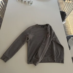 Grå tröja från Henri Lloyd - Säljer en stilren grå tröja från Henri Lloyd med ett litet broderat emblem på armen. Perfekt för en avslappnad stil. Den är i storlek m men passar s. 