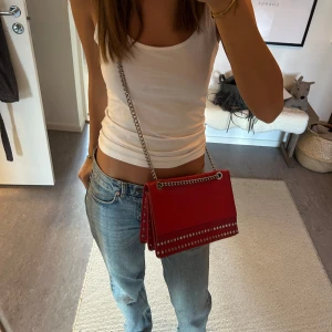 Röd axelväska med kedja - Snygg röd axelväska med silverfärgad kedja och nitar. Perfekt för att ge en färgklick till din outfit. Väskan har en stilren design med detaljer som ger en edgy känsla.