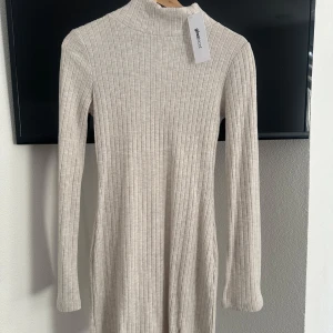 Beige ribbad klänning från Gina Tricot - Säljer en oanvänd stilren beige ribbad klänning från Gina Tricot. Klänningen har långa ärmar och en liten slits framtill. Perfekt för en chic och bekväm look. Passar bra till både vardag och fest!