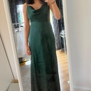 Mörkgrön balklänning - Elegant mörkgrön långklänning med en stilren design. Klänningen har en ärmlös stil och en smickrande passform som framhäver figuren. Dragkedja där bak och spännbara axelband. 